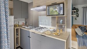 A compact kitchen with a gas stove, a microwave, and a sink. There are cabinets and a small shelf with kitchen items. - Proche Lac & Océan | Piscine | Camping 4* (Hourtin)