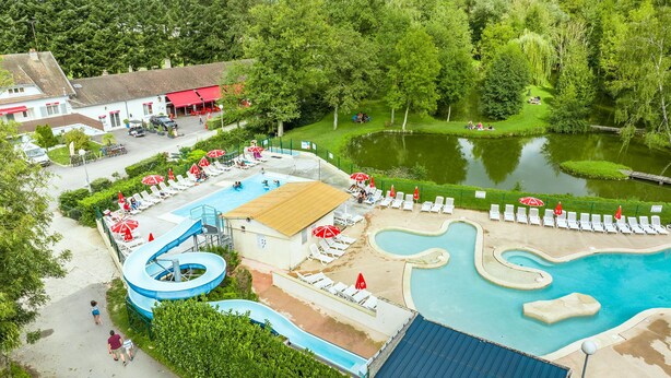 A swimming pool with a slide, surrounded by lounge chairs and umbrellas, with a forested area in the background.