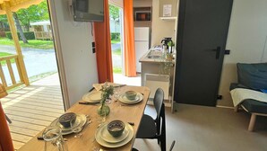 A dining area with a wooden table set for two, a television mounted on the wall, and a kitchen area with a microwave and refrigerator.