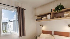 A bedroom with a bed, a window with curtains, a bedside table, a shelf with books and a plant, and a headboard.