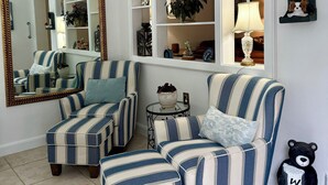 Una sala de estar con sillones a rayas, otomanas, un espejo, estantes con objetos decorativos y un letrero de bienvenida con forma de oso.