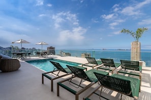Un área de piscina en la azotea con sillones de descanso y sombrillas, con vistas a un horizonte urbano y al mar.
