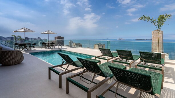 A rooftop pool area with lounge chairs and umbrellas, overlooking a cityscape and the sea. - (1006) Apartamento com 2 Suítes e Vista para o Cristo (Rio de Janeiro)