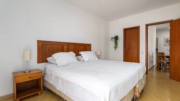 Quarto com cabeceira de madeira, uma cama de solteiro com roupa de cama branca, mesa de cabeceira com abajur e uma pequena prateleira.