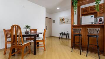 Uma sala de jantar com mesa e cadeiras de madeira, uma pequena cozinha com geladeira e pia, e uma planta pendurada.