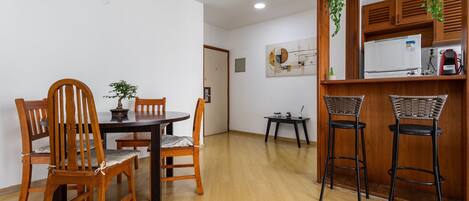 A dining area with a wooden table and chairs, a small kitchen area with a refrigerator and a sink, and a hanging plant.