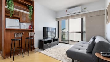 Uma sala de estar moderna com um sofá cinzento, uma mesa de centro, uma televisão e uma área de refeições com frigorífico, micro-ondas e banquetas.