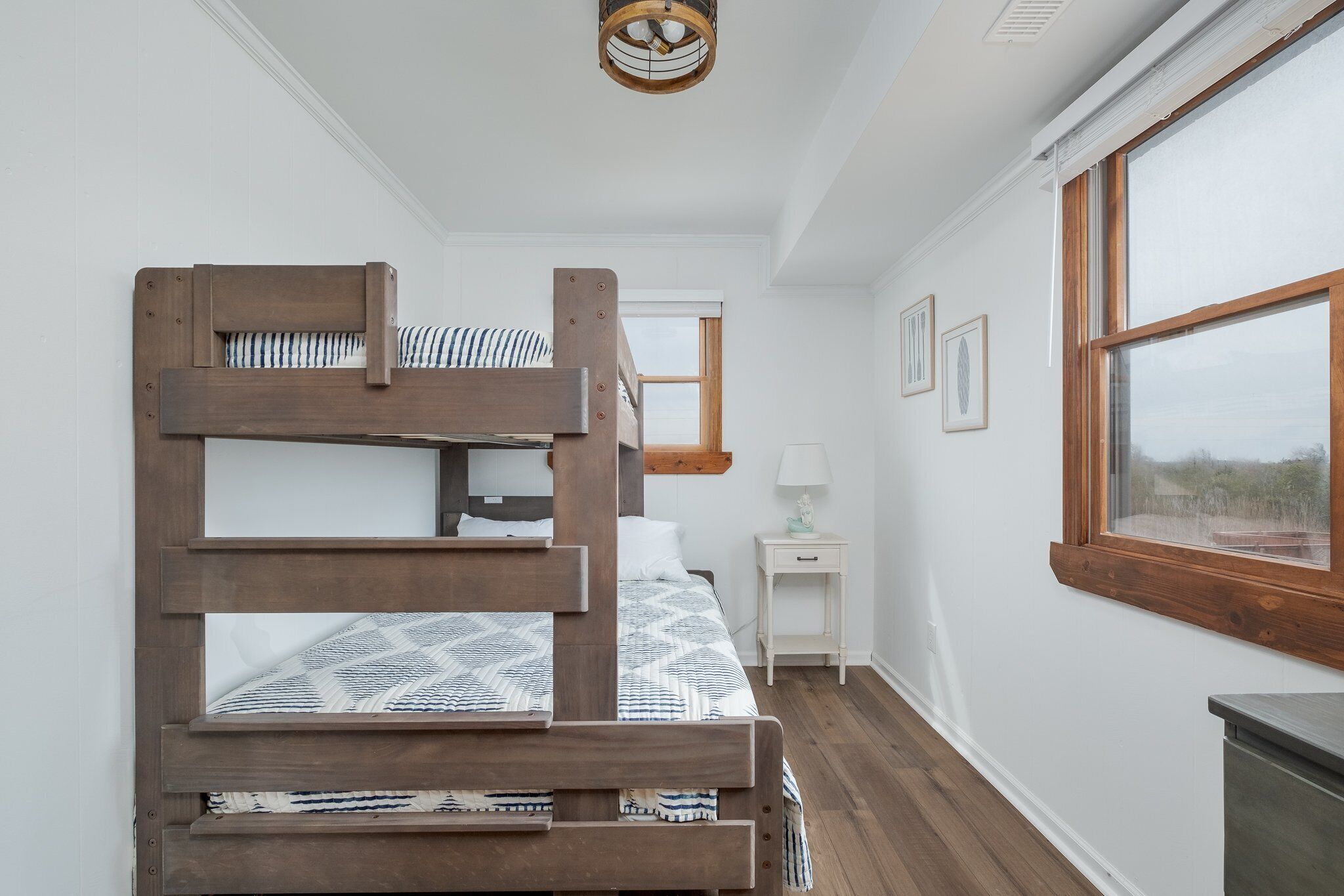 Uma cama de solteiro com estrutura de madeira e roupa de cama estampada, uma pequena mesa de cabeceira com abajur e uma janela com vista para o exterior.