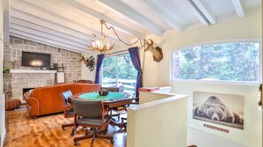 Uma sala de estar acolhedora com lareira, televisão fixada acima dela, mesa de jantar com cadeiras e uma grande janela com vista para o exterior.
