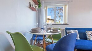 A dining area with a round table set for four, surrounded by vibrant green and blue chairs. A blue sofa is visible in the background, and a window with a view of a building and trees is on the wall. - 1bed-sleeps3 Parking-nearcitycentre-jlr-nec (Birmingham)