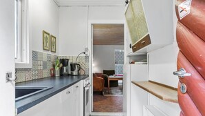 A narrow kitchen with white cabinets, a black countertop, and a patterned backsplash. There is a coffee maker, a kettle, and a small table with a chair. A door with a red panel and a handle is visible on the right. - 2 bedroom amazing home in Kolind (Kolind)