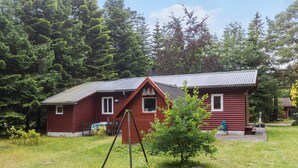 A red wooden cabin with a metal roof, surrounded by a grassy yard and dense forest. - 2 bedroom amazing home in Kolind (Kolind)
