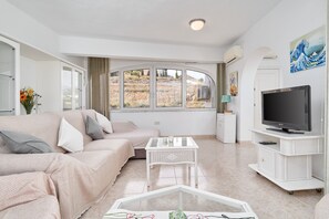 Una sala de estar con un sofá, una mesa de centro de vidrio, un televisor sobre un mueble bajo y un amplio ventanal con vistas a una colina.