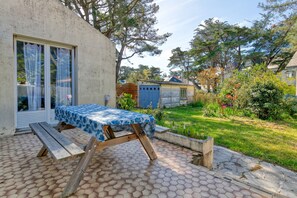 Une terrasse avec une table et des bancs en bois, un espace couvert avec une table, et un jardin agrémenté de diverses plantes et d’une clôture.