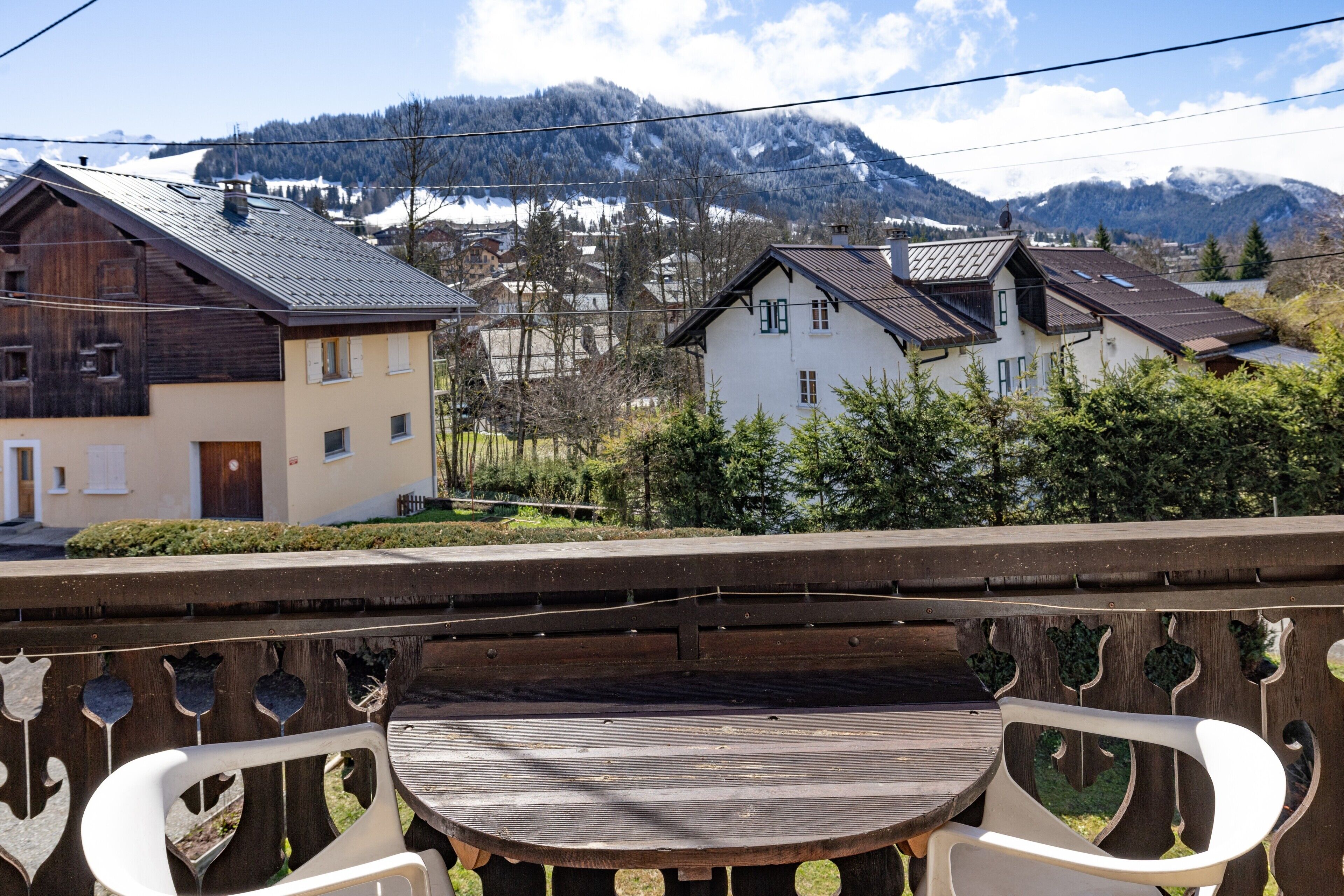 Un balcón con una mesa y sillas de madera, con vista a un pueblo con casas y montañas al fondo.