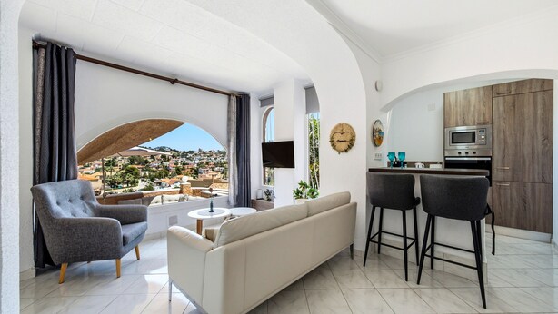 A modern living room with a sofa, armchair, and bar stools, a kitchen with a microwave, and a view of a cityscape through an arched window. - Holiday Home 'Brandy' with Sea View, Wi-Fi and Air Conditioning (Calpe)