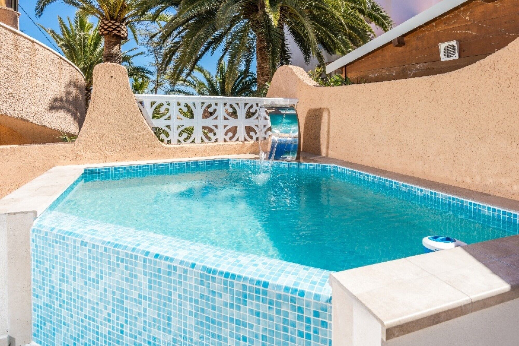 Una piscina con agua cristalina azul, rodeada de muros color arena y palmeras.