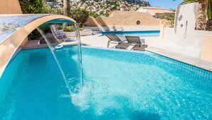 A swimming pool with a waterfall feature, lounge chairs, and a view of a hillside village. - Holiday Home 'Brandy' with Sea View, Wi-Fi and Air Conditioning (Calpe)