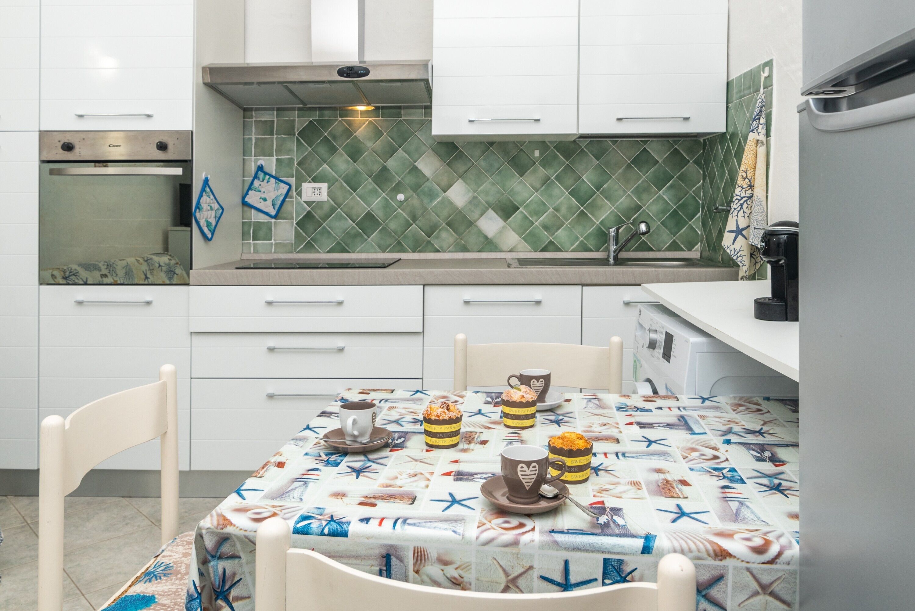 Une cuisine avec des meubles blancs, un pare-feu en carreaux et une table dressée avec une nappe aux motifs marins.