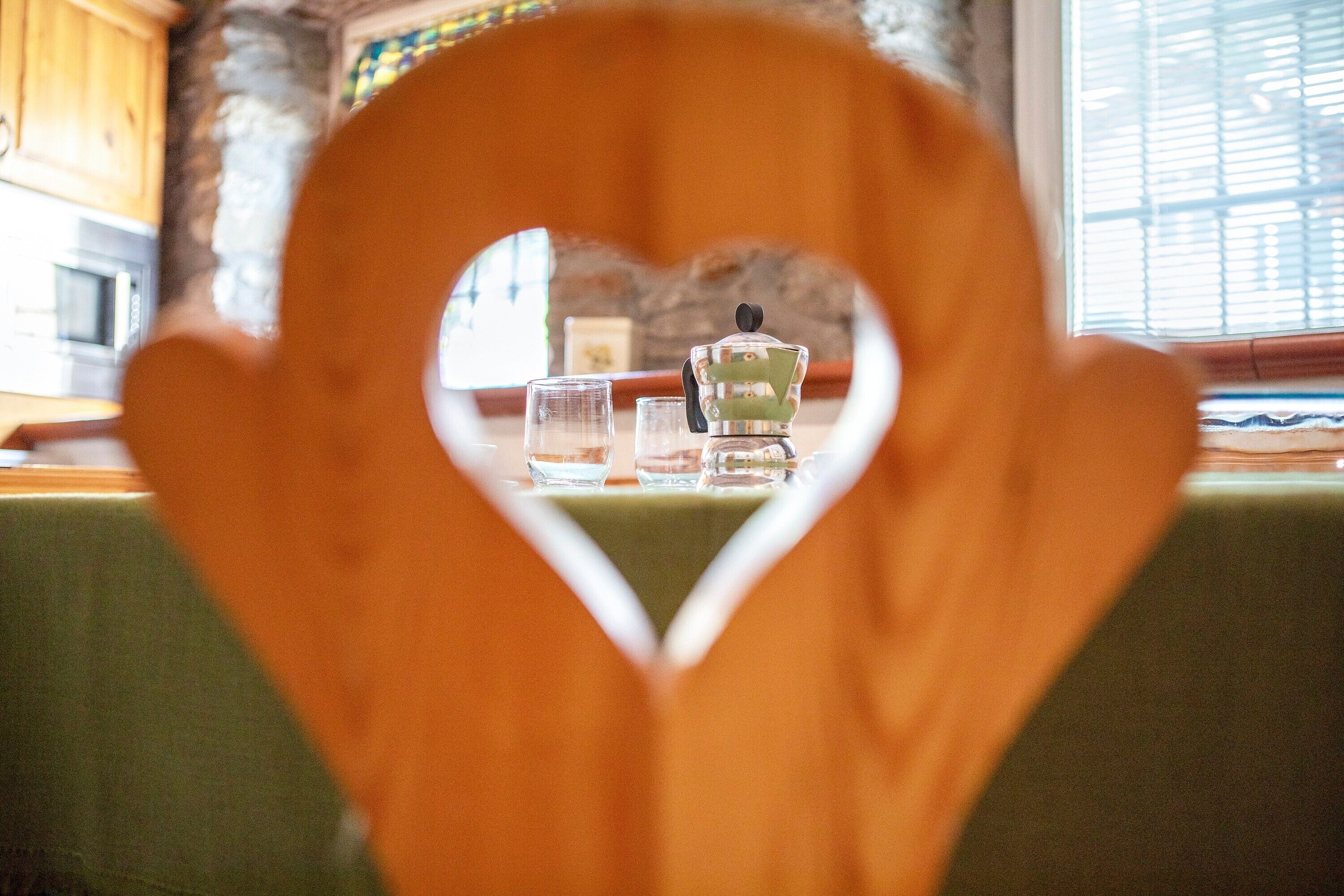 A heart-shaped cutout framing a table with a teapot and glasses, a window with blinds, and a stone wall.
