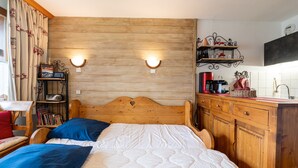 Ein Bett mit hölzernem Kopfteil, zwei Nachttischlampen, eine Kommode mit Schubladen und ein kleines Regal mit Büchern und einem Radio.