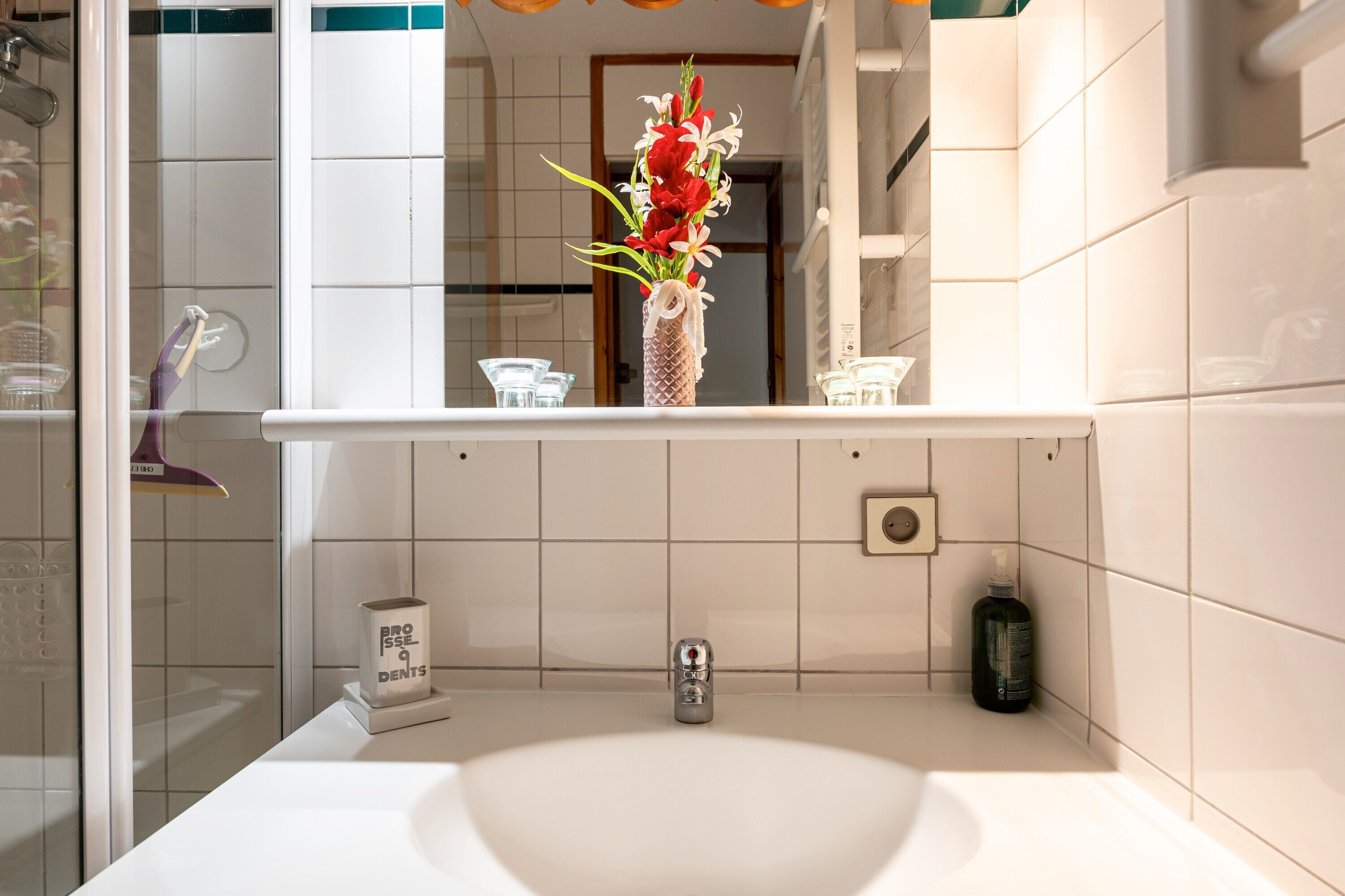A bathroom with a white sink, a mirror, a vase with flowers, a toothbrush holder, and a soap dispenser.