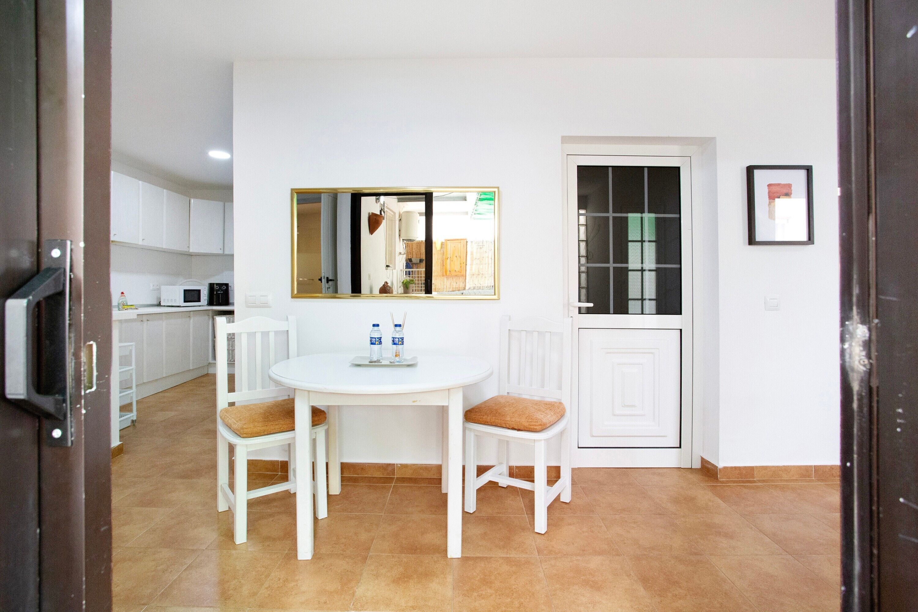 Une cuisine compacte avec des meubles blancs, une table à manger blanche accompagnée de deux chaises, et une fenêtre donnant sur l’extérieur.