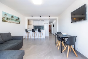 A modern living area with a sofa, dining table with chairs, a kitchen, and a TV mounted on the wall.