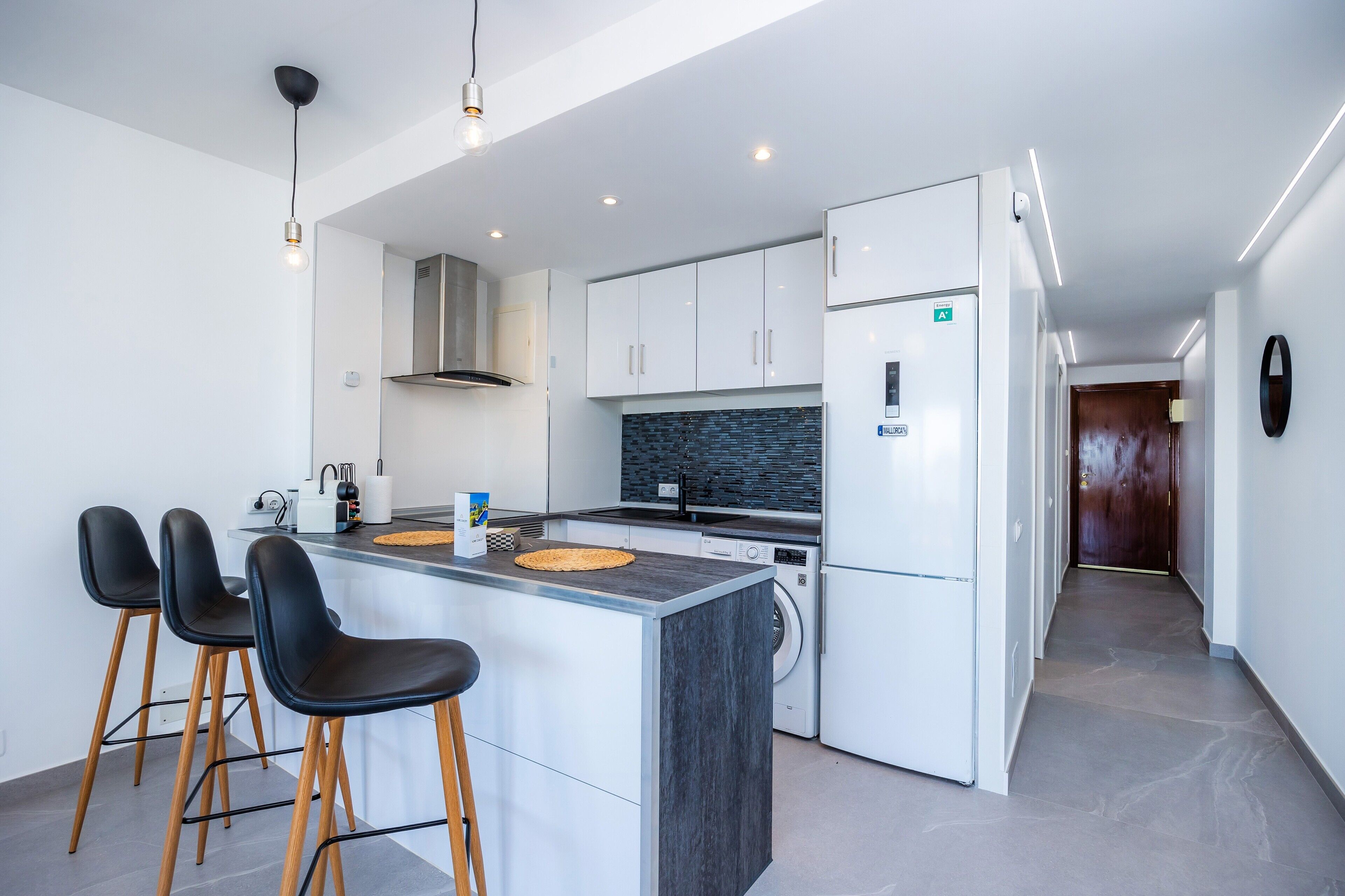 A modern kitchen with a central island, bar stools, a washing machine, and a hallway leading to a door.
