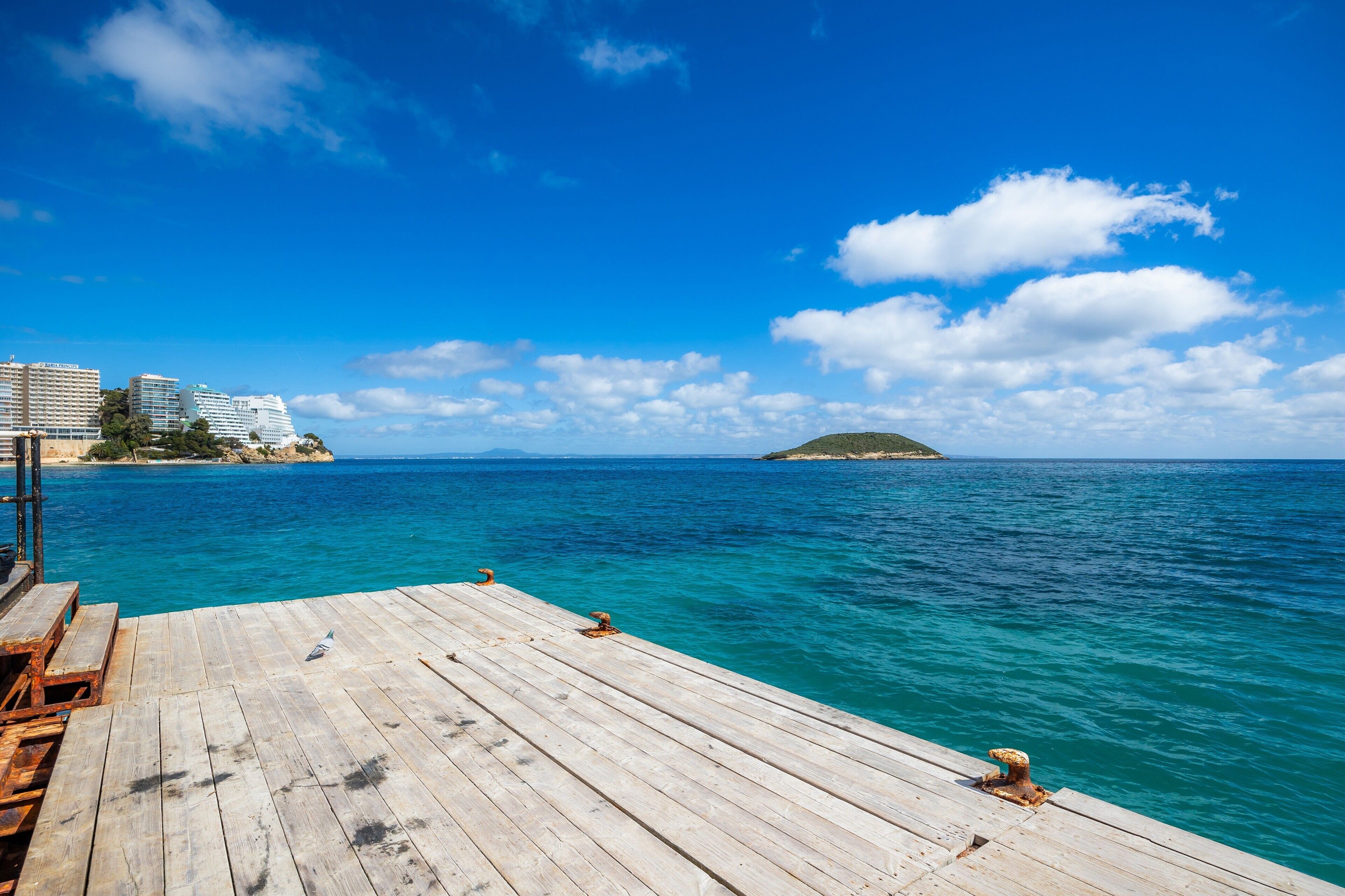 Eine Holzstege, der sich in ein klares, blaues Meer erstreckt, mit einer entfernten Insel und Gebäuden an der Küste.