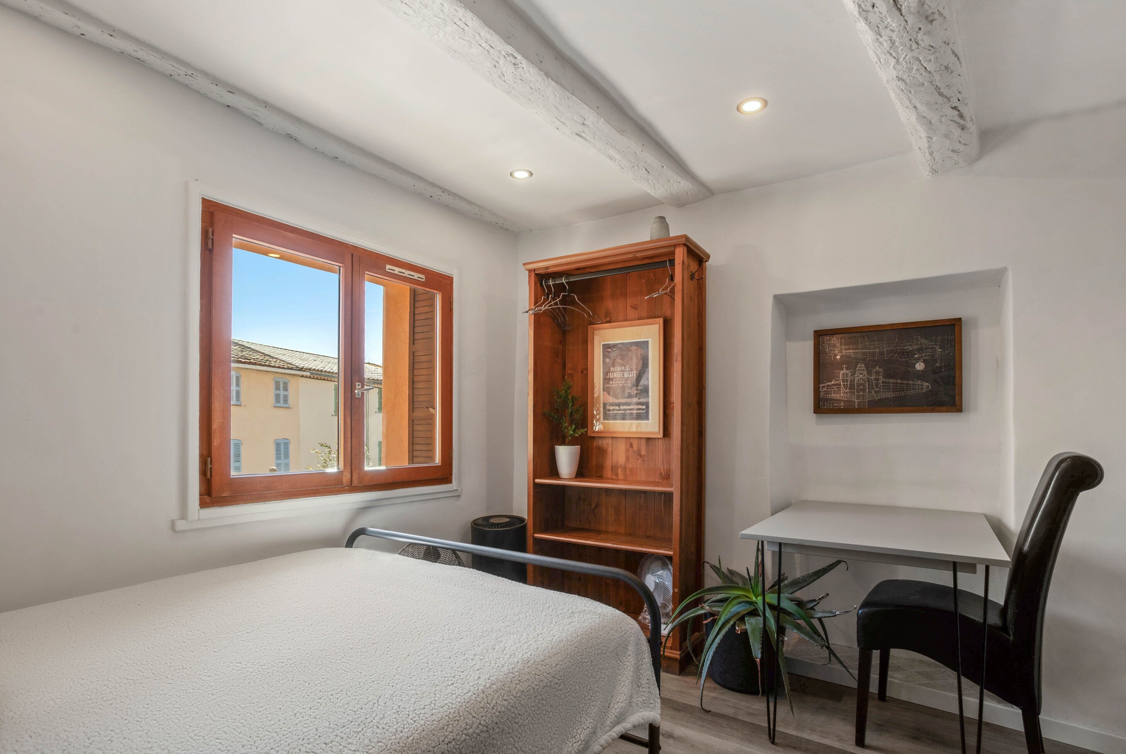 Une chambre à coucher avec un lit, une étagère en bois ornée d’une plante, un bureau avec une chaise et un tableau encadré accroché au mur.