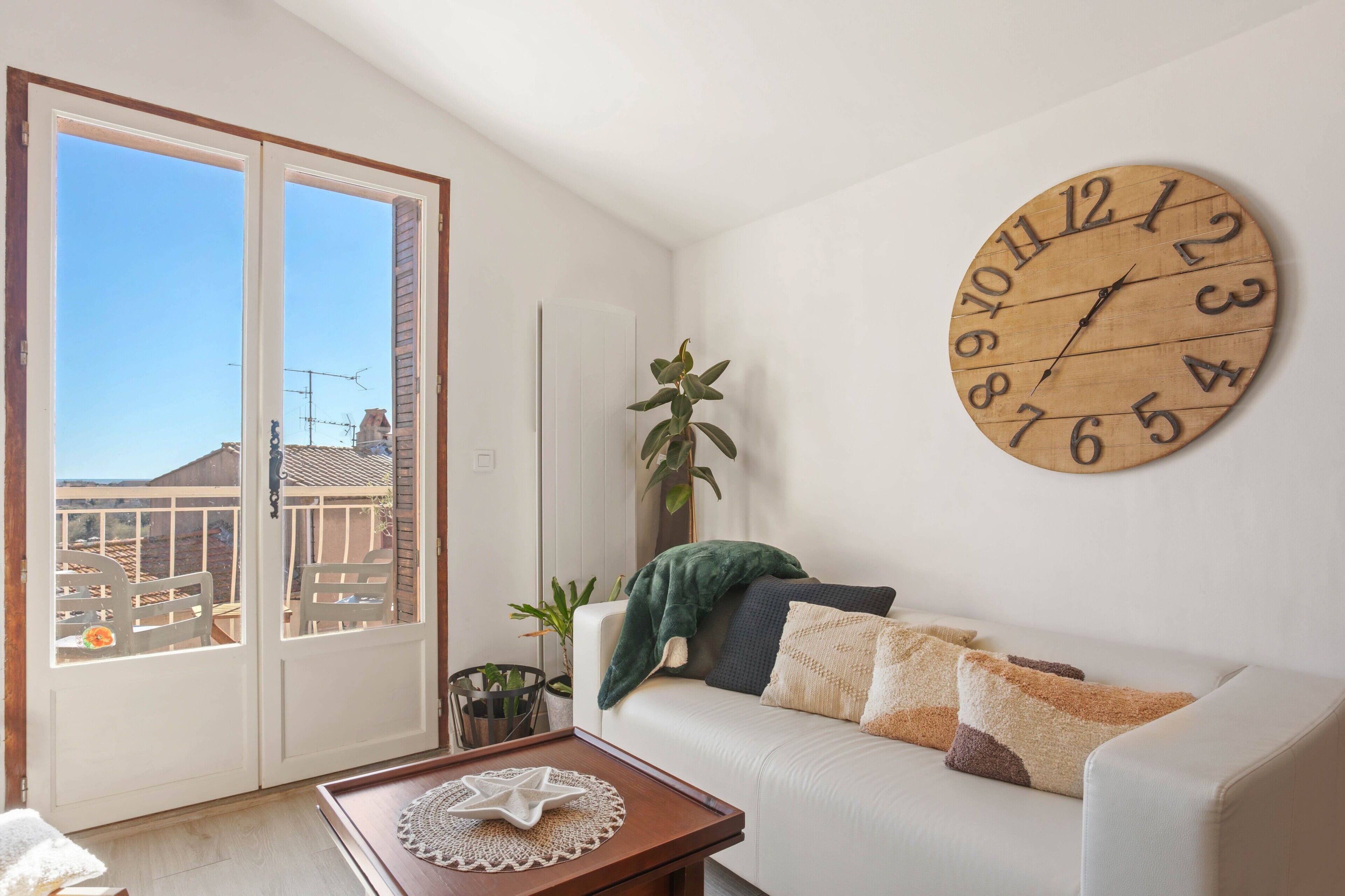 Un salon avec une grande horloge en bois, un canapé avec des coussins, une petite table sur laquelle se trouve un objet en forme d’étoile, et une vue à travers une porte vitrée.
