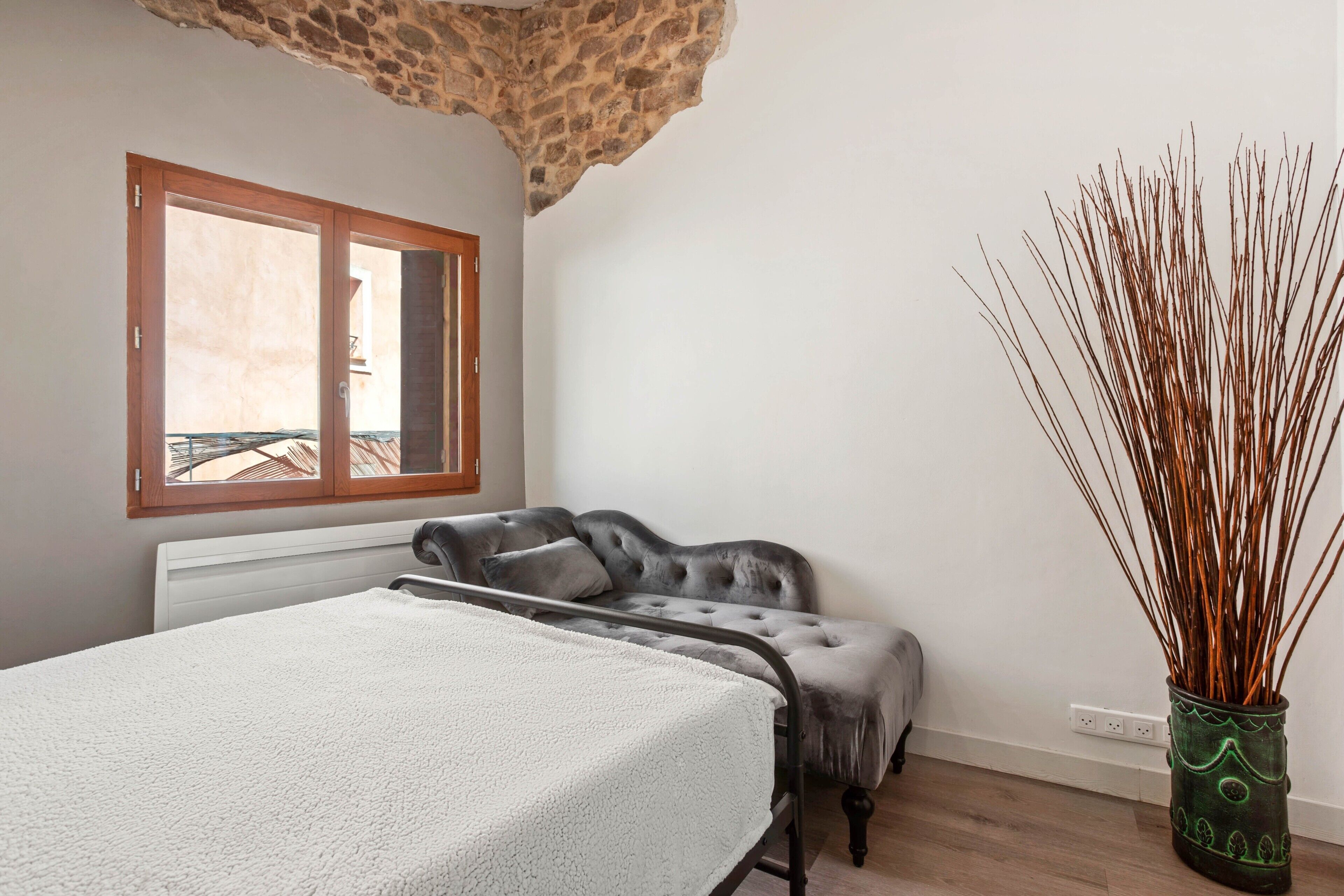 Een slaapkamer met een bed, een grijze bank, een stenen plafond, een raam en een groene vaas met takken.