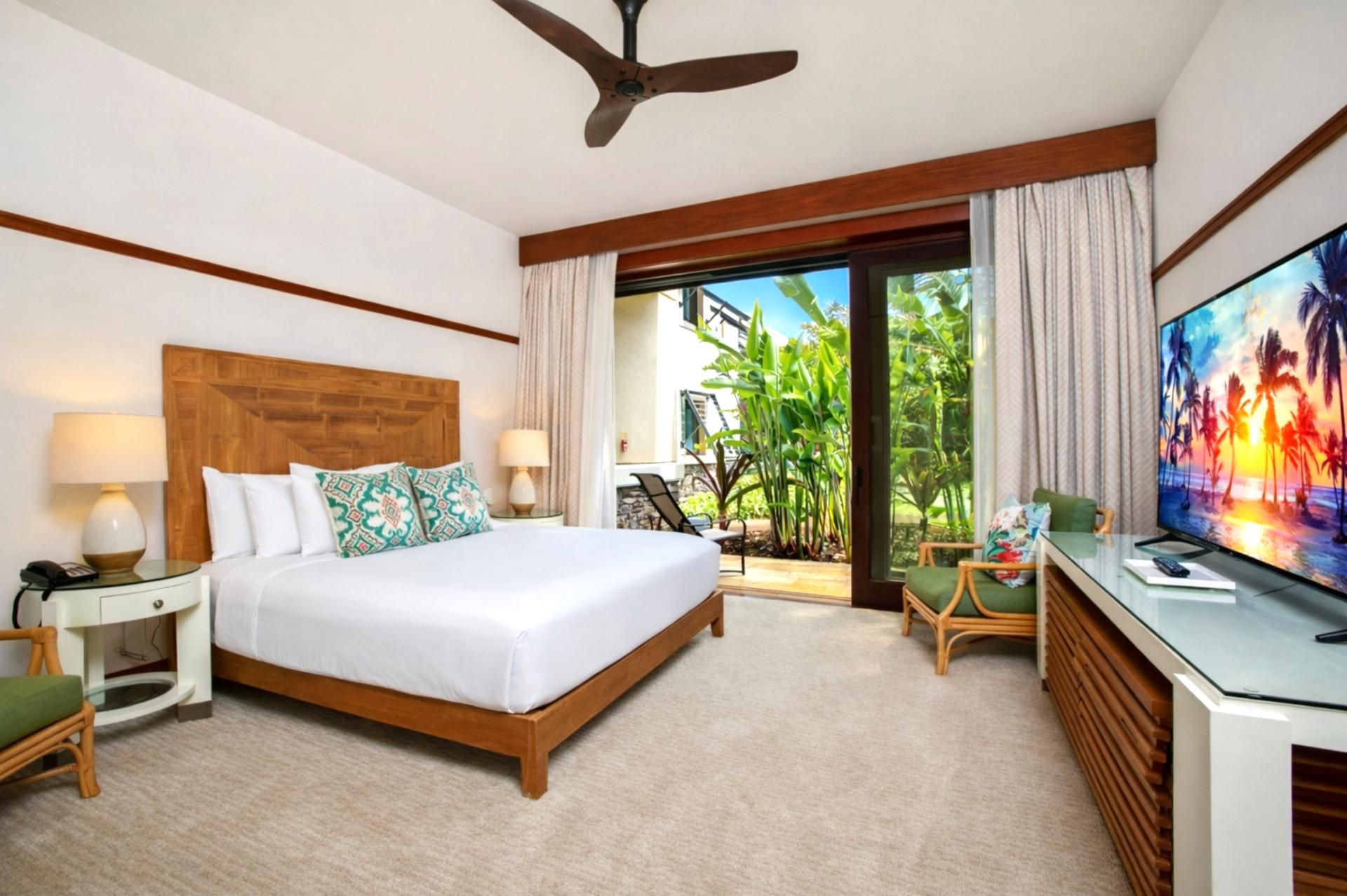 A hotel room with a bed, a desk with a TV, a ceiling fan, and a view of greenery outside.