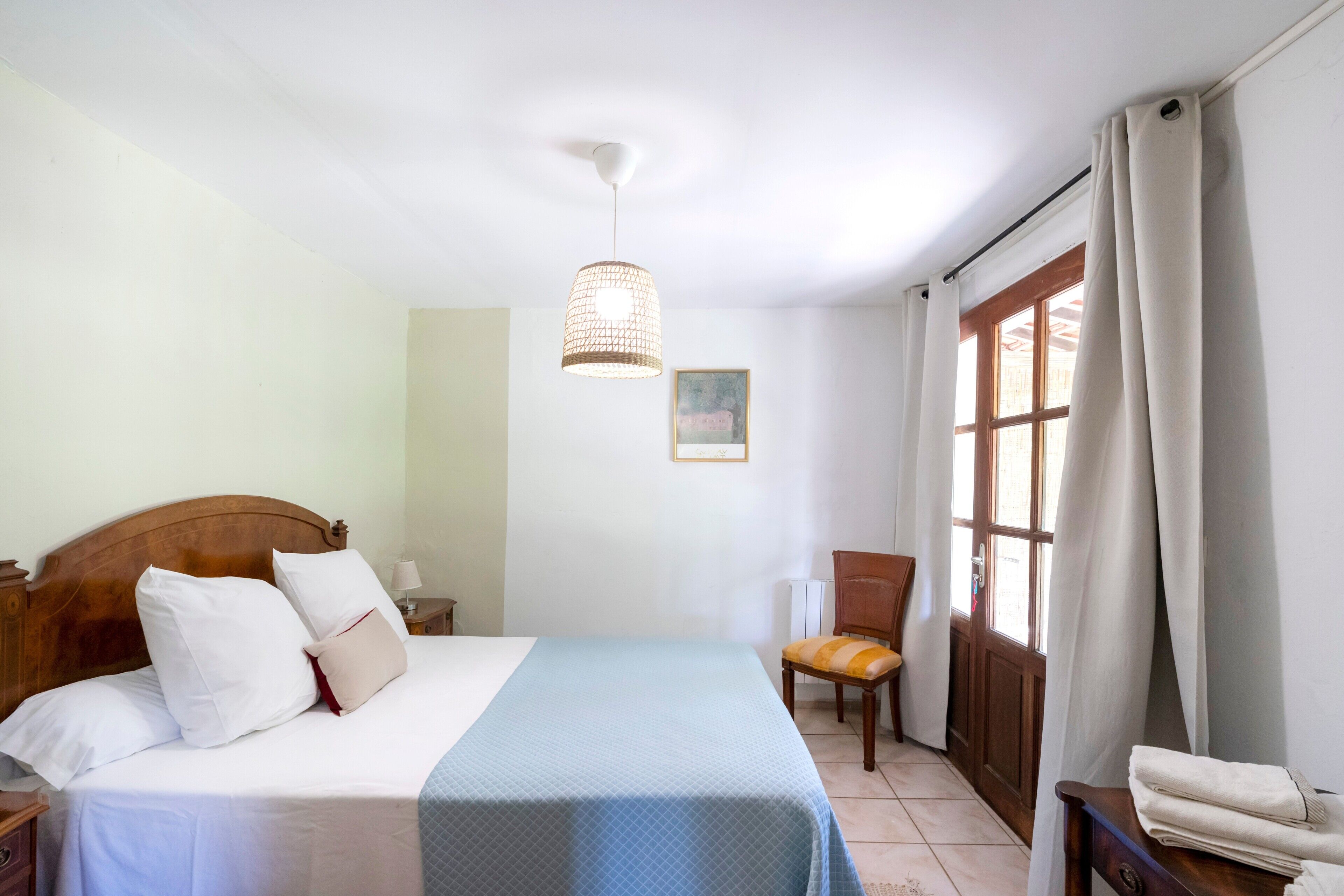 Een slaapkamer met een houten bed, een nachtkastje, een stoel, een raam met gordijnen en een hanglamp.