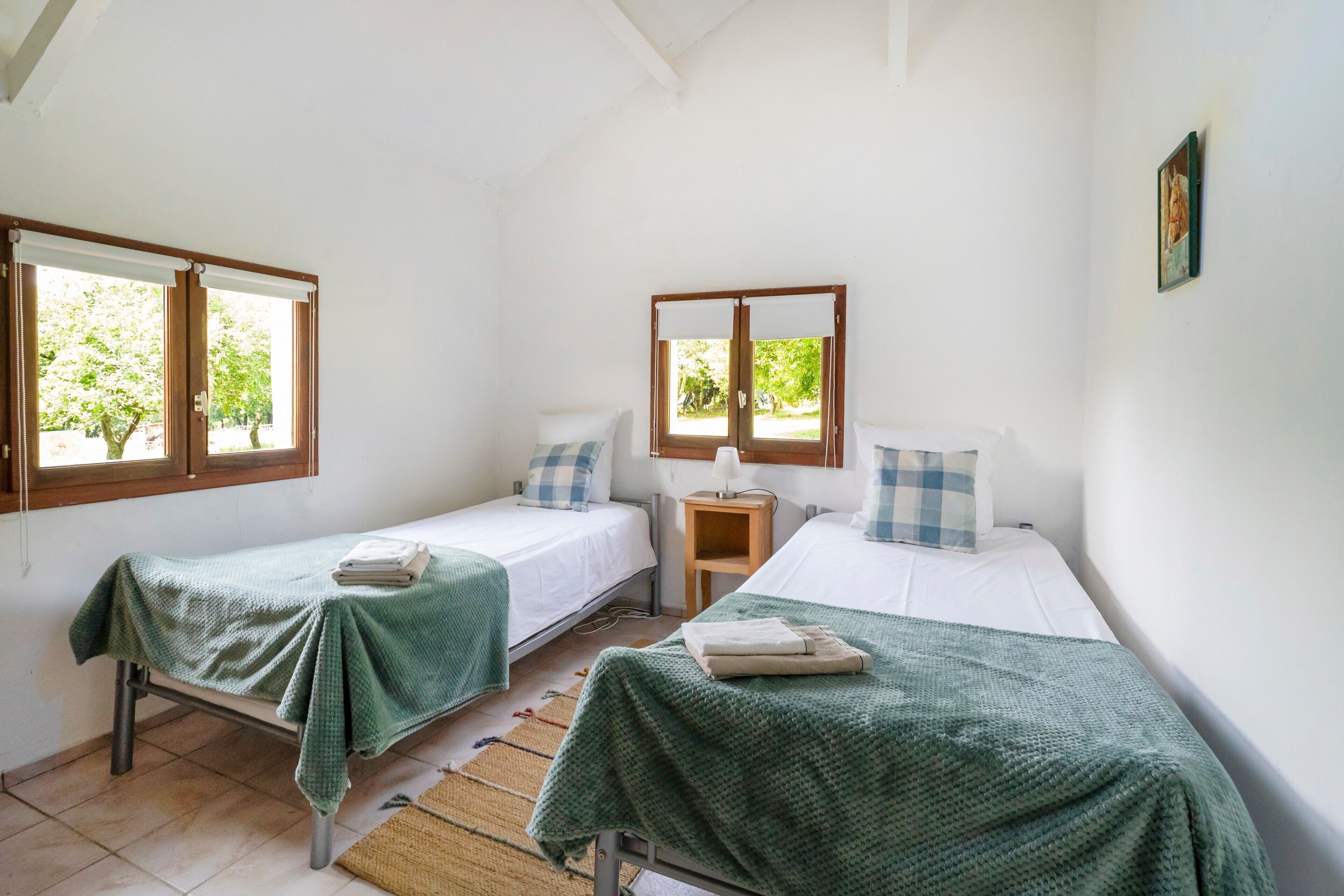 A room with two single beds, a small bedside table with a lamp, and a window with a view of greenery.