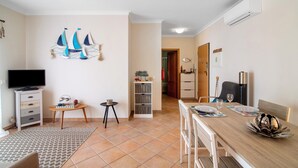 Un salon avec un téléviseur, une table à manger dressée pour deux personnes, une bibliothèque et une porte donnant sur une autre pièce.