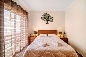 A neatly made bed with a tree wall art, two bedside tables with lamps, and a window with curtains.