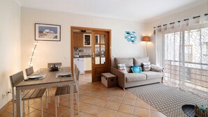 Un coin repas avec une table et des chaises, un canapé avec des coussins, un tapis à motifs, une cuisine en arrière-plan et une grande fenêtre avec des rideaux.