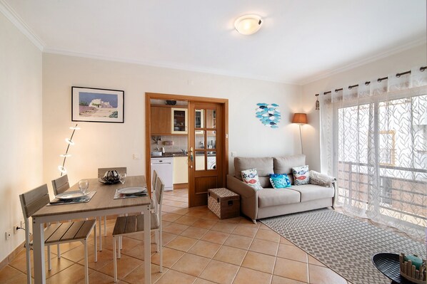 A dining area with a table and chairs, a sofa with cushions, a patterned rug, a kitchen in the background, and a large window with curtains.