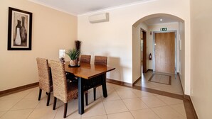 A dining area with a wooden table and woven chairs, a framed artwork on the wall, and an air conditioning unit.