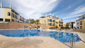 A swimming pool with a stone wall, a clear blue sky, and multi-story buildings with balconies.