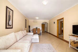 Una sala de estar con un sofá modular, un televisor sobre un mueble bajo y un comedor al fondo.