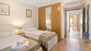 A hotel room with two beds, a wardrobe, a mirror, a bedside table with a lamp, and a potted plant.