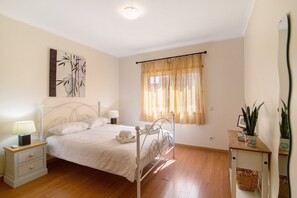 Un dormitorio con cama, mesitas de noche, una lámpara, ventana con cortinas, una obra de arte en la pared y una planta en maceta.