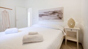 A neatly made bed with two towels, a white nightstand with a decorative lamp, and a painting on the wall.