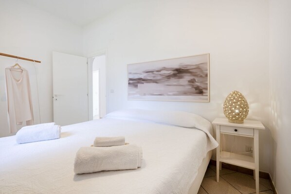 Una cama bien tendida con dos toallas, una mesita de noche blanca con una lámpara decorativa y un cuadro en la pared.
