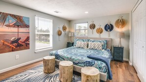 Ein Schlafzimmer mit einem großen Bett, hölzernen Hocker, einem Wandbild mit Strandmotiv und Blick ins Freie durch das Fenster.