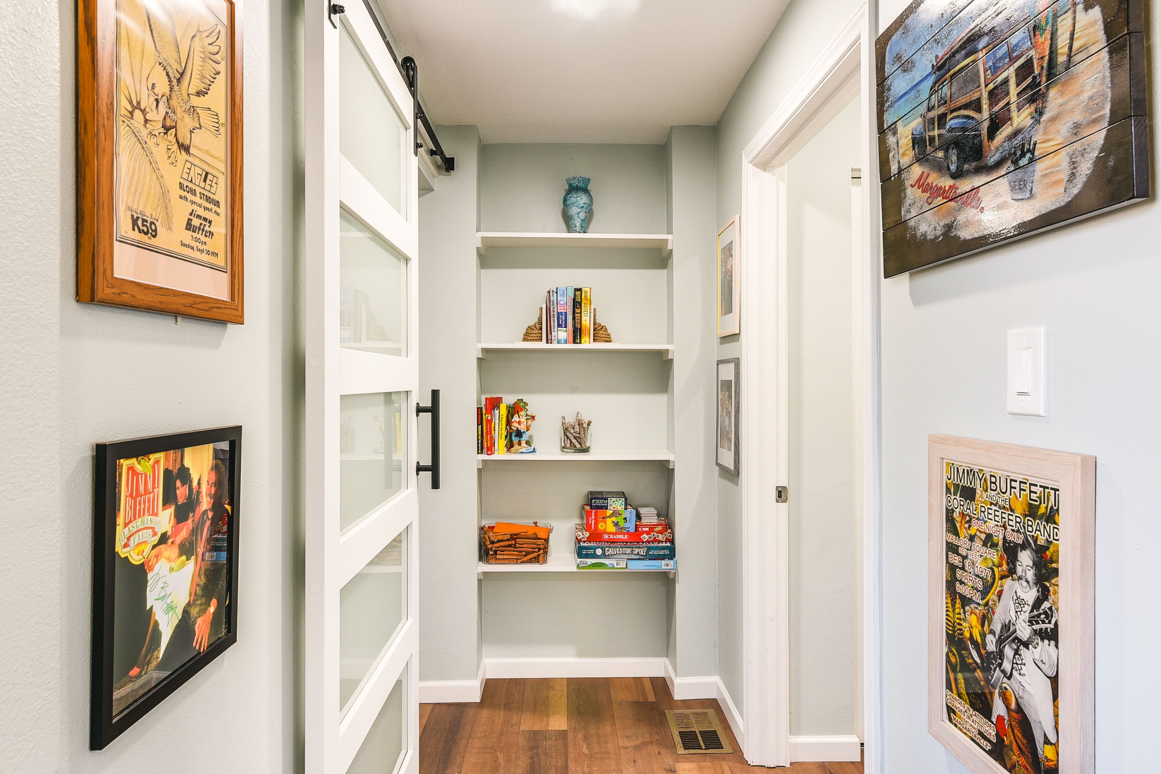 A narrow hallway with white walls, wooden flooring, and a central white door. Shelves on either side hold books and decorative items.