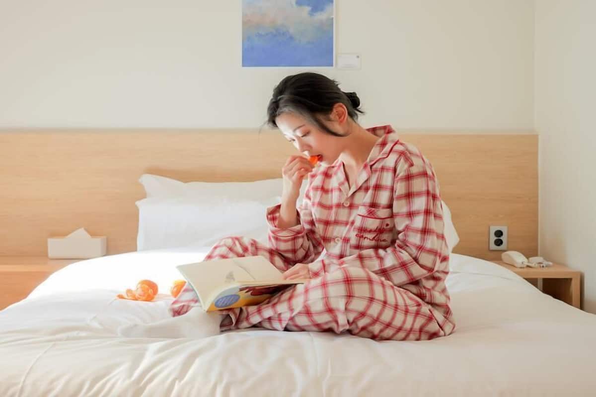 Une personne assise sur un lit dans une chambre d’hôtel, en train de lire un livre.